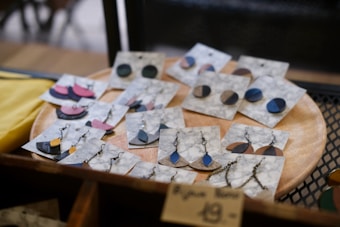 A collection of handmade earrings is displayed on marble-patterned cards, resting on a wooden circular tray. The earrings come in various shapes and colors, including blue, black, and pink, with both stud and dangling designs featuring metal chains and hooks. The display gives a rustic and artisanal feel.