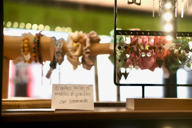 A display of colorful handmade bracelets and dangling earrings is set on a wooden stand. The bracelets are made of various beads, while the earrings are intricate and decorative. There is a small sign in French indicating the availability of different sizes and models.