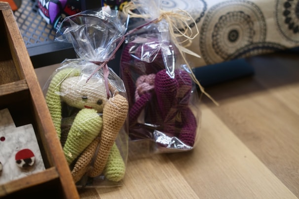 Two crocheted toys wrapped in clear plastic are placed on a wooden surface. One toy is green and beige, while the other is purple and pink. Both are shaped like dolls with elongated limbs. There is a wooden shelf in the corner with a few other items displayed, including a small, white, and red round object. In the background, a fabric with black and white circular patterns is visible.