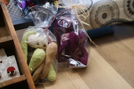 Two crocheted toys wrapped in clear plastic are placed on a wooden surface. One toy is green and beige, while the other is purple and pink. Both are shaped like dolls with elongated limbs. There is a wooden shelf in the corner with a few other items displayed, including a small, white, and red round object. In the background, a fabric with black and white circular patterns is visible.