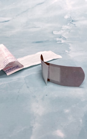 A bandage is placed on a light blue surface. One bandage is still in its packaging, partially open, and the other is unwrapped with the adhesive side visible.
