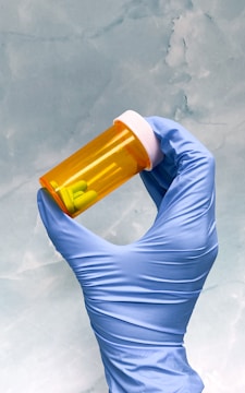 A gloved hand holds a small orange prescription bottle filled with yellow tablets. The background is a smooth, light blue texture.