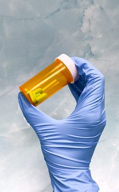 A gloved hand holds a small orange prescription bottle filled with yellow tablets. The background is a smooth, light blue texture.