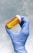 A gloved hand holds a small orange prescription bottle filled with yellow tablets. The background is a smooth, light blue texture.