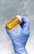 A gloved hand holds a small orange prescription bottle filled with yellow tablets. The background is a smooth, light blue texture.