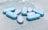 A collection of blue and light purple pills is scattered on a soft, textured surface. The pills have imprints on them, indicating they may be pharmaceutical tablets. The lighting is bright, highlighting the smooth texture of the pills.