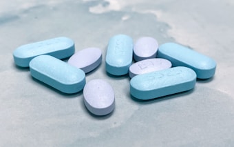 A collection of blue and light purple pills is scattered on a soft, textured surface. The pills have imprints on them, indicating they may be pharmaceutical tablets. The lighting is bright, highlighting the smooth texture of the pills.