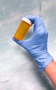 A hand wearing a blue medical glove is holding an empty orange prescription bottle with a white cap against a light blue marbled background.