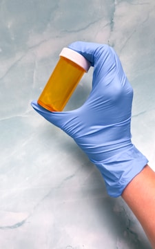 A hand wearing a blue medical glove is holding an empty orange prescription bottle with a white cap against a light blue marbled background.