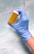 A hand wearing a blue medical glove is holding an empty orange prescription bottle with a white cap against a light blue marbled background.