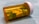A translucent orange prescription pill bottle lies on its side, containing a mix of yellow and green tablets. The tablets are marked with numbers and letters. The white cap is securely attached, and the background is a soft, blurred blue.