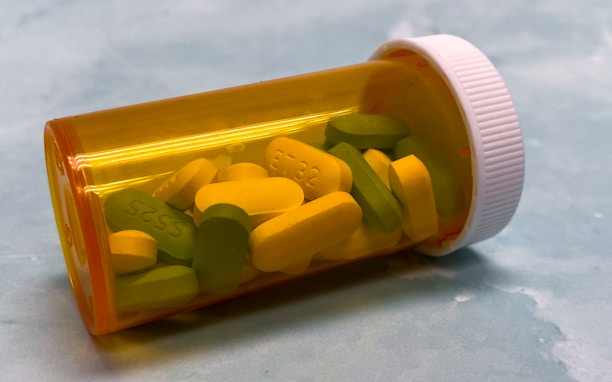 A translucent orange prescription pill bottle lies on its side, containing a mix of yellow and green tablets. The tablets are marked with numbers and letters. The white cap is securely attached, and the background is a soft, blurred blue.