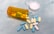 A variety of colorful pills are scattered on a light blue surface, with additional tablets contained inside an orange prescription bottle lying on its side. The white cap of the bottle is placed nearby.
