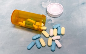 A variety of colorful pills are scattered on a light blue surface, with additional tablets contained inside an orange prescription bottle lying on its side. The white cap of the bottle is placed nearby.