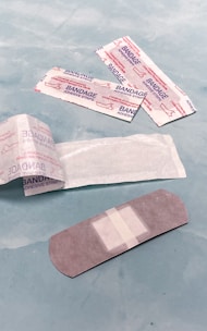 Several adhesive bandages and their packaging are placed on a light blue surface. The bandages are surrounded by opened and unopened wrappers that display the word 'BANDAGE' in red.