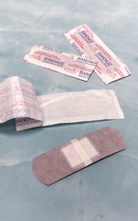 Several adhesive bandages and their packaging are placed on a light blue surface. The bandages are surrounded by opened and unopened wrappers that display the word 'BANDAGE' in red.