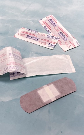 Several adhesive bandages and their packaging are placed on a light blue surface. The bandages are surrounded by opened and unopened wrappers that display the word 'BANDAGE' in red.