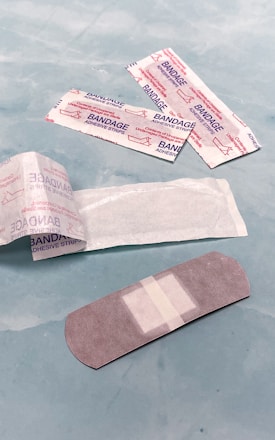 Several adhesive bandages and their packaging are placed on a light blue surface. The bandages are surrounded by opened and unopened wrappers that display the word 'BANDAGE' in red.