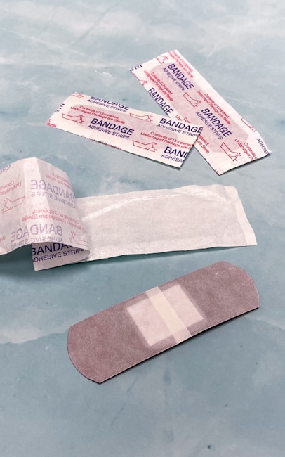Several adhesive bandages and their packaging are placed on a light blue surface. The bandages are surrounded by opened and unopened wrappers that display the word 'BANDAGE' in red.