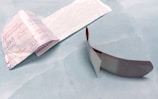 An opened adhesive bandage is lying on a light blue surface. The bandage is partially unwrapped, revealing a white pad on one side. The wrapper features text and branding details. Next to the bandage is the paper backing that has been removed.