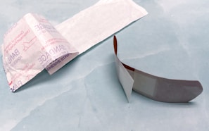 An opened adhesive bandage is lying on a light blue surface. The bandage is partially unwrapped, revealing a white pad on one side. The wrapper features text and branding details. Next to the bandage is the paper backing that has been removed.