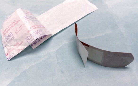 An opened adhesive bandage is lying on a light blue surface. The bandage is partially unwrapped, revealing a white pad on one side. The wrapper features text and branding details. Next to the bandage is the paper backing that has been removed.