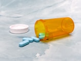 A transparent orange pill bottle lies on its side on a light blue, textured surface. The white cap is off to the side, and several light blue, oval-shaped tablets have spilled out onto the surface.