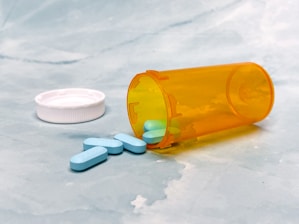 A transparent orange pill bottle lies on its side on a light blue, textured surface. The white cap is off to the side, and several light blue, oval-shaped tablets have spilled out onto the surface.