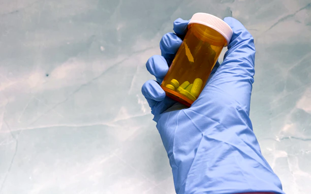 A close-up of a pharmacist's hands holding prescription medication bottles against a soft blue background.