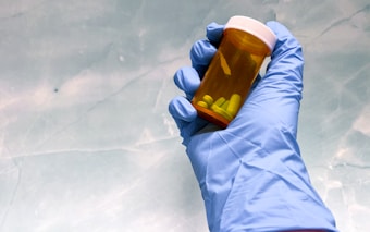 A hand wearing a blue latex glove holds a small amber prescription bottle containing yellow pills. The background is a soft, light blue surface, possibly a counter or tabletop.