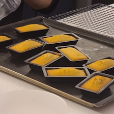 Several small rectangular cakes, golden brown in color, are neatly placed on a baking tray. The cakes are still in their baking molds, and there is a cooling rack in the background. Part of a person's hand can be seen near the corner of the tray, indicating handling or preparation.