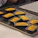 Several small rectangular cakes, golden brown in color, are neatly placed on a baking tray. The cakes are still in their baking molds, and there is a cooling rack in the background. Part of a person's hand can be seen near the corner of the tray, indicating handling or preparation.