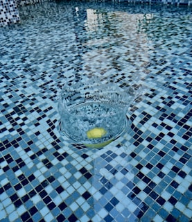 A tiled swimming pool with a colorful mosaic pattern. A splash in the water creates ripples around a yellow object that seems to have just entered the water.