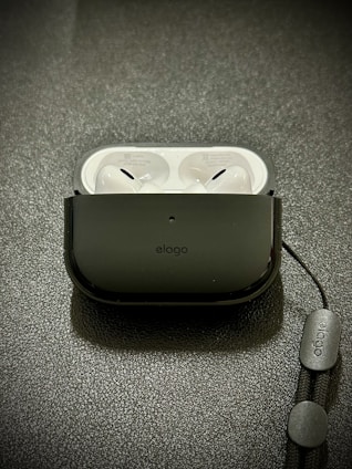 A pair of white wireless earbuds resting inside a black protective case on a textured surface. A lanyard is attached to the case, featuring branding text.