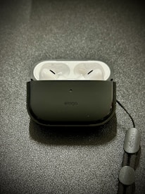 A pair of white wireless earbuds resting inside a black protective case on a textured surface. A lanyard is attached to the case, featuring branding text.