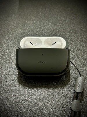 A pair of white wireless earbuds resting inside a black protective case on a textured surface. A lanyard is attached to the case, featuring branding text.