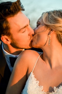 A couple sharing an intimate kiss, captured in warm, golden lighting. The woman is wearing an elegant white dress with lace detailing and an elaborate earring. The man is dressed in a dark suit with a bow tie. Their faces are closely touching, conveying a sense of love and affection.