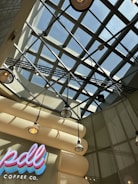 The image features a modern interior ceiling design with a grid-like glass structure allowing natural light to filter through. Beneath the ceiling, several hanging industrial-style pendant lights add to the ambiance. The wall displays a bright, colorful sign with 'pdl coffee co.' written in neon-like letters.