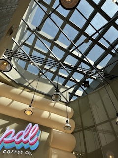 The image features a modern interior ceiling design with a grid-like glass structure allowing natural light to filter through. Beneath the ceiling, several hanging industrial-style pendant lights add to the ambiance. The wall displays a bright, colorful sign with 'pdl coffee co.' written in neon-like letters.