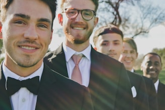 A group of five men dressed in formal black suits with white shirts and ties or bow ties, standing in a row outdoors. They are smiling, and the sun casts a warm light, creating lens flares across the image. A blurred tree is in the background, suggesting a natural or park setting.