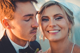 A close-up of a couple smiling warmly. The woman has light blonde hair styled neatly with long earrings, and her eyes are wide open with a joyful expression. The man, dressed in a black suit, has his eyes gently closed, leaning towards her with a content expression.