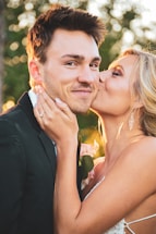 A couple is depicted with a woman kissing a smiling man on the cheek. The man is wearing a black suit, while the woman is in a white dress. Her hand, adorned with a ring, gently holds his face. There is a soft, natural background with blurred greenery.