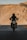 A silhouetted motorcyclist rides down a road with barren, rugged hills in the background. The motorcycle's headlight is on, creating a striking contrast against the shadow. The scene conveys a sense of adventure and solitude.