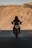 A silhouetted motorcyclist rides down a road with barren, rugged hills in the background. The motorcycle's headlight is on, creating a striking contrast against the shadow. The scene conveys a sense of adventure and solitude.