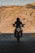 A silhouetted motorcyclist rides down a road with barren, rugged hills in the background. The motorcycle's headlight is on, creating a striking contrast against the shadow. The scene conveys a sense of adventure and solitude.