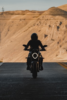 A silhouetted motorcyclist rides down a road with barren, rugged hills in the background. The motorcycle's headlight is on, creating a striking contrast against the shadow. The scene conveys a sense of adventure and solitude.