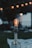 A vintage light bulb is lit in an outdoor setting, resting on a decorative lamp base. In the blurred background, several small, warm-toned lights create a bokeh effect against the dusk sky.