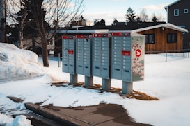 Several clustered mailboxes are standing in a snowy residential area. The ground is covered in snow, with some patches of exposed pathway. A wooden house and a fence can be seen in the background. The mailboxes have red accents and one features decorative designs.