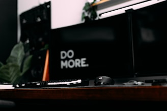 A modern and minimalistic workspace with a dark theme features a keyboard, computer mouse, and dual monitors. The words 'DO MORE' are prominently displayed on one of the screens, suggesting a motivational message.