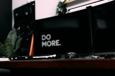 A modern and minimalistic workspace with a dark theme features a keyboard, computer mouse, and dual monitors. The words 'DO MORE' are prominently displayed on one of the screens, suggesting a motivational message.